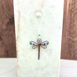 Small copper wire wrapped and beaded hanging dragonfly ornament. it's hanging from a suction cup to a marble slab in front of a wooden backdrop
