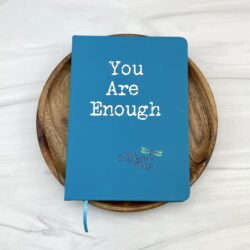 Turquoise journal notebook with phrase You Are Enough and On A Dragonfly's Wings logo on cover laying on a wooden dish on a marble counter top