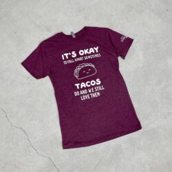 Burgundy tee shirt laying flat on a cement background with the saying “It’s okay to fall apart sometimes tacos do and we still love them.” and a picture of a smiling taco on the front in white print