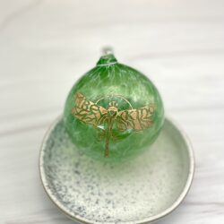 Green blown glass bulb ornament with a gold etched dragonfly and the words Be Kind incorporated in the wings on a white and grey ceramic plate on a white marble counter