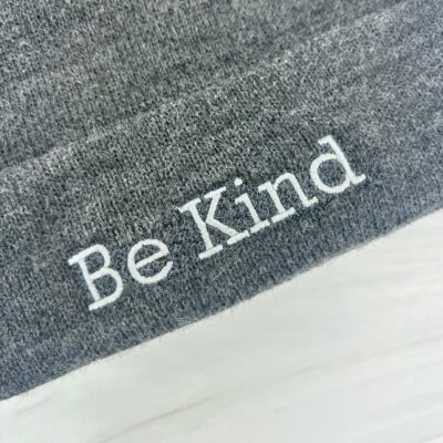 Dark grey knit beanie with "Be Kind" embroidered in light grey on brim laying flat on marble trivet