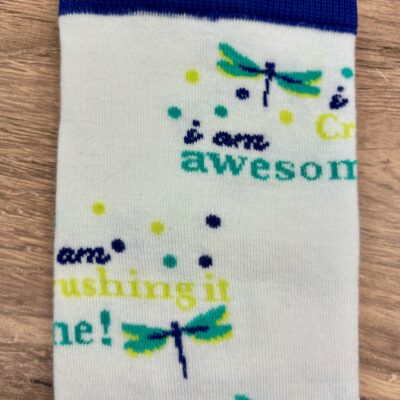 Close up of one knit sock showing the dragonfly and phrases "i am awesome" and "i am crushing it" on wooden countertop background