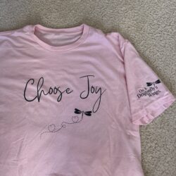 Light pink tee shirt laying flat on a carpet background with the saying “Choose Joy” and a picture of the On A Dragonfly's Wings dragonfly in black print