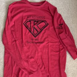 Red long-sleeved tee shirt laying flat on a carpet background with the saying “Kindness is my Superpower” and a monogram K in black print