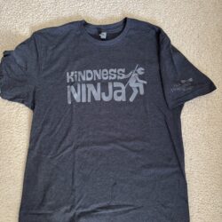 Navy heather tee shirt laying flat on a carpet background with the saying “KiNDNESS NINJA” and a picture of a cartoon ninja in distressed white print