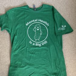 Green tee shirt laying flat on a carpet background with the saying “Mental Health is a Big Dill” and a picture of a cartoon pickle smiling in white print